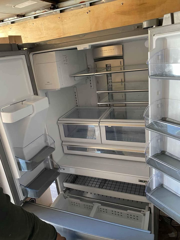 An empty white refrigerator with its doors open, showing the interior shelves, door bins, and lower drawer. A person's arm is visible at the bottom left corner. condition new