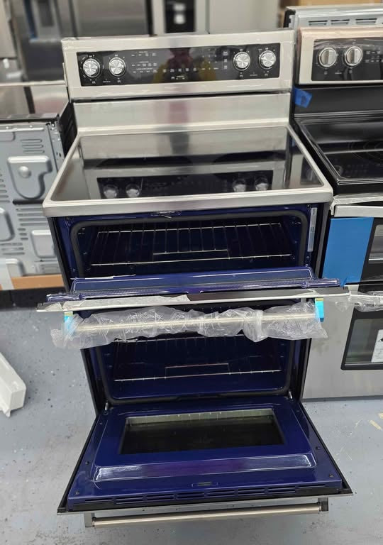 A freestanding electric range with a stainless steel exterior, black glass cooktop, and dark blue interior oven chamber. The oven door is open, showing the interior racks and glass door, and the appliance is photographed in what appears to be a showroom or warehouse setting with other appliances in the background.