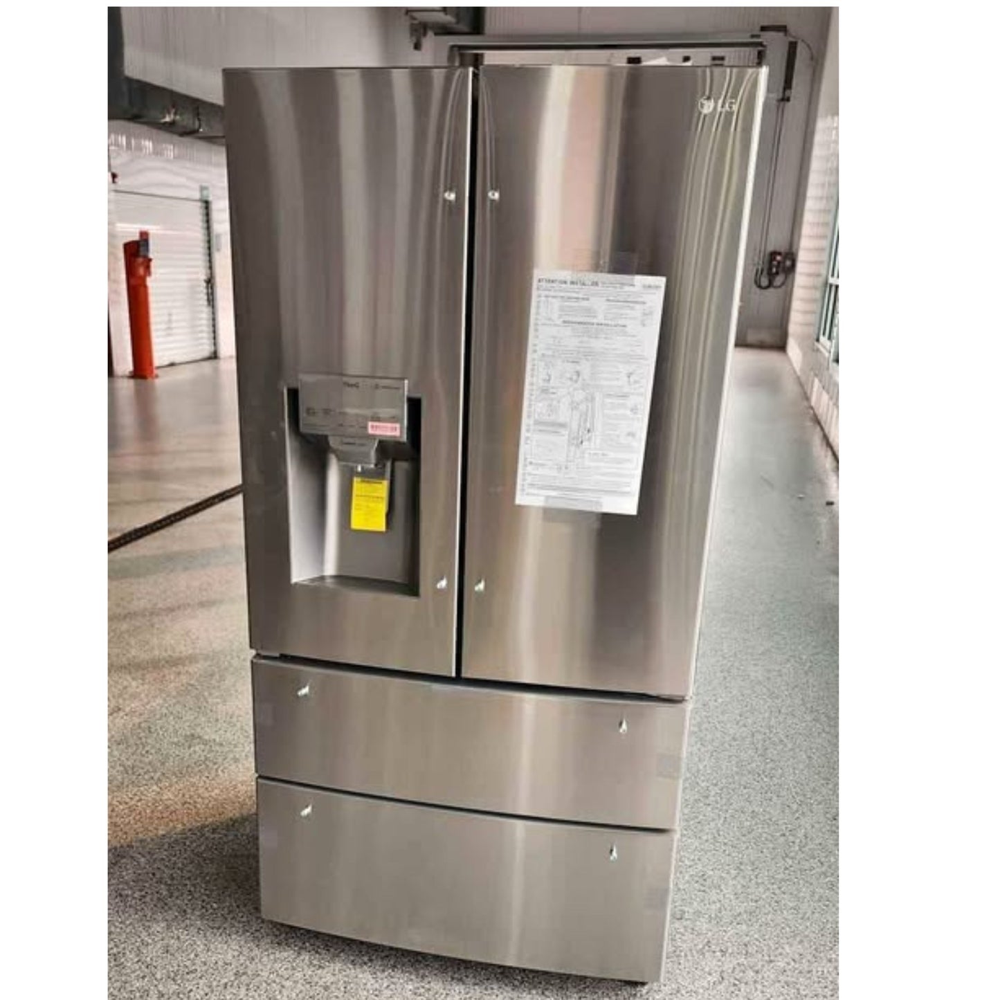 A stainless steel French door refrigerator with a bottom freezer drawer, standing in an indoor showroom or warehouse area. A white paper sheet is attached to the right door.
