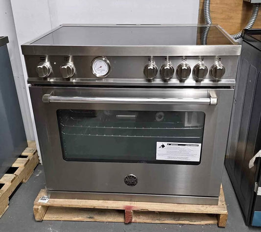 A stainless steel freestanding range oven with a glass door, control knobs along the top, and a wood pallet underneath, photographed in a warehouse setting.