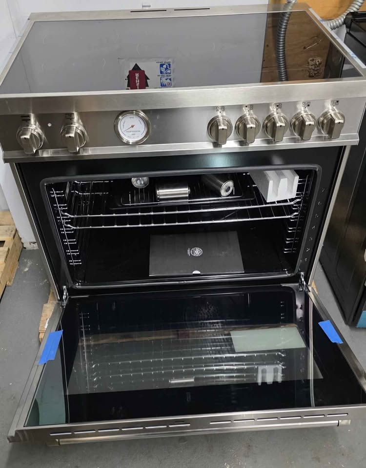 A stainless steel freestanding oven with its door open, showing the interior racks and glass door. The front has multiple control knobs, a round dial gauge, and a smooth cooktop surface.