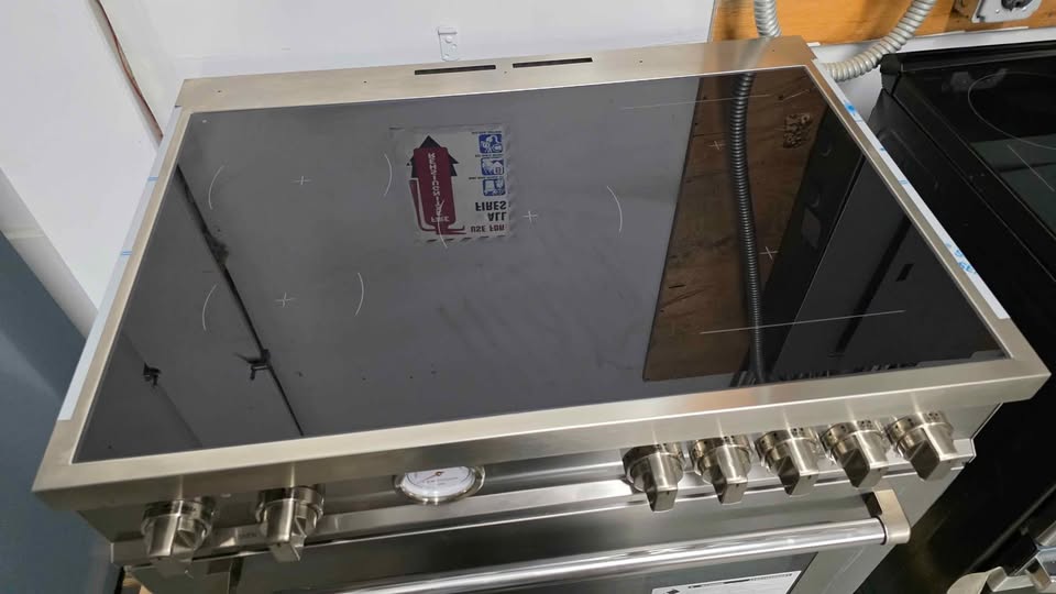 A stainless steel gas range with a black glass cooktop and multiple control knobs on the front, photographed in what appears to be a workshop or showroom.