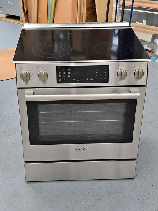 A Bosch freestanding electric range with a stainless steel body, black glass cooktop, five control knobs, and a digital control panel, photographed in what appears to be a warehouse or showroom setting.
