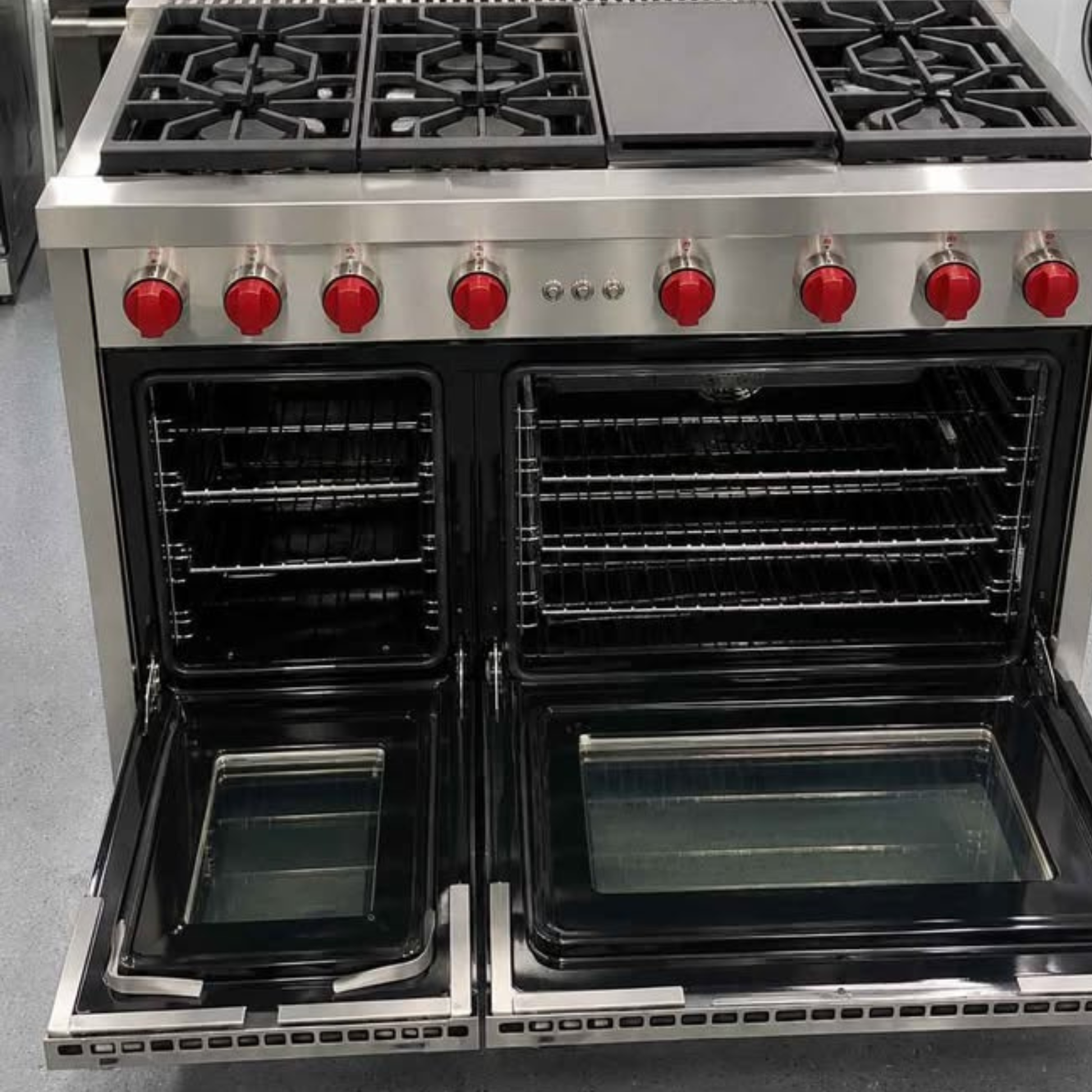 A large stainless steel freestanding gas range with two open ovens, black grates, a smooth cooktop, and bright red control knobs, photographed in a showroom setting.