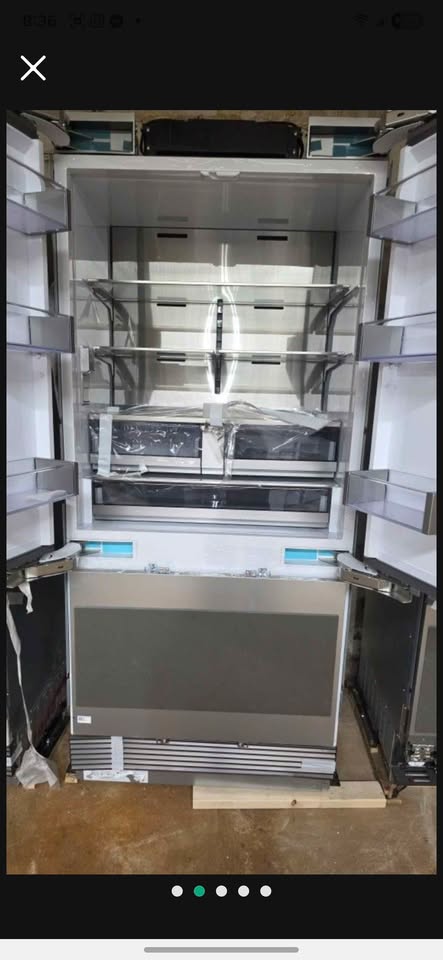 An open stainless steel refrigerator or freezer with multiple glass shelves and a dark lower drawer, photographed in what appears to be a warehouse or storage area with a concrete floor.