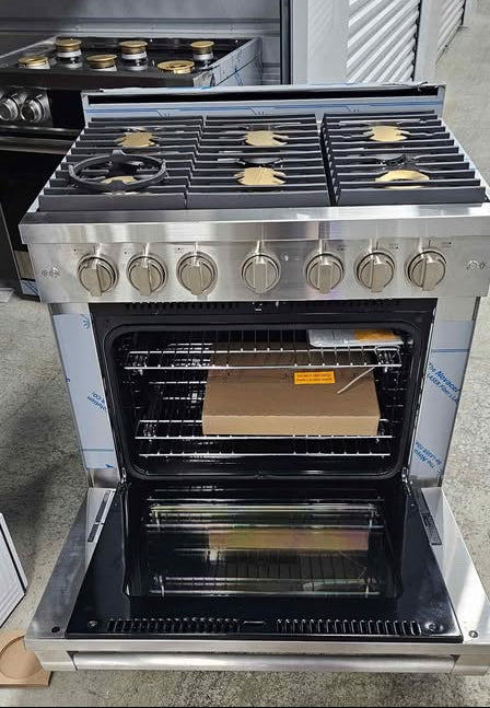 A stainless steel freestanding gas range stove with an open oven door, shown in a showroom. The cooktop has six burners with black grates and silver control knobs.