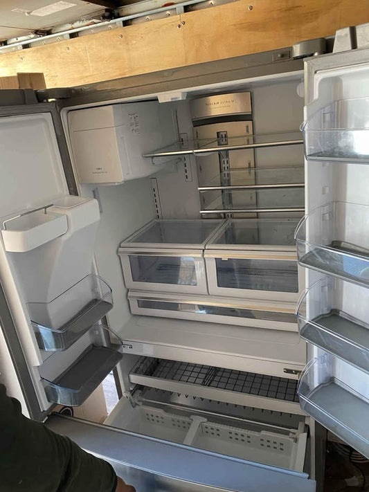 An empty white refrigerator with its doors open, showing the interior shelves, door bins, and lower drawer. A person's arm is visible at the bottom left corner. condition new
