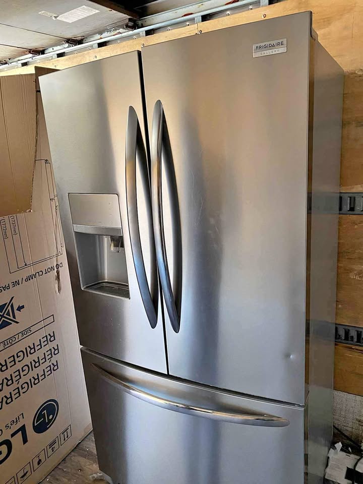 A stainless steel Frigidore French door refrigerator with a bottom freezer drawer, standing next to a cardboard shipping box in a warehouse or showroom setting.