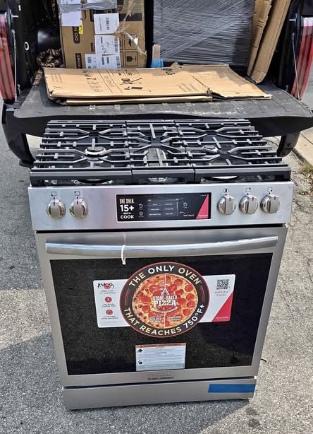 A stainless steel freestanding gas range with a black glass oven door, front control panel, and a large circular 'We Only Overheat' sticker, photographed outdoors on a concrete surface.