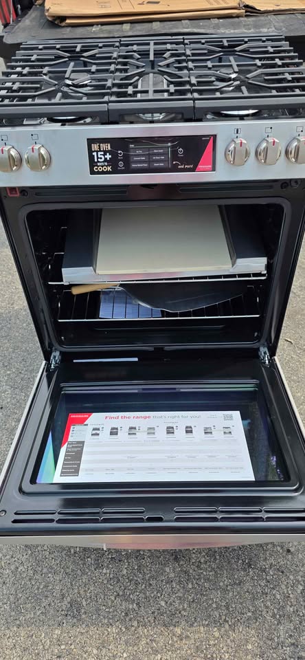 A freestanding gas range stove with an open oven door, showing the interior racks and a control panel with knobs and a digital display. A product manual is visible inside the oven door tray.