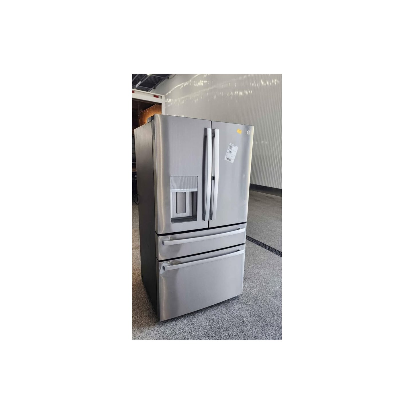 A stainless steel French door refrigerator with a bottom freezer drawer, shown in a warehouse or showroom setting with a gray floor and light gray walls.