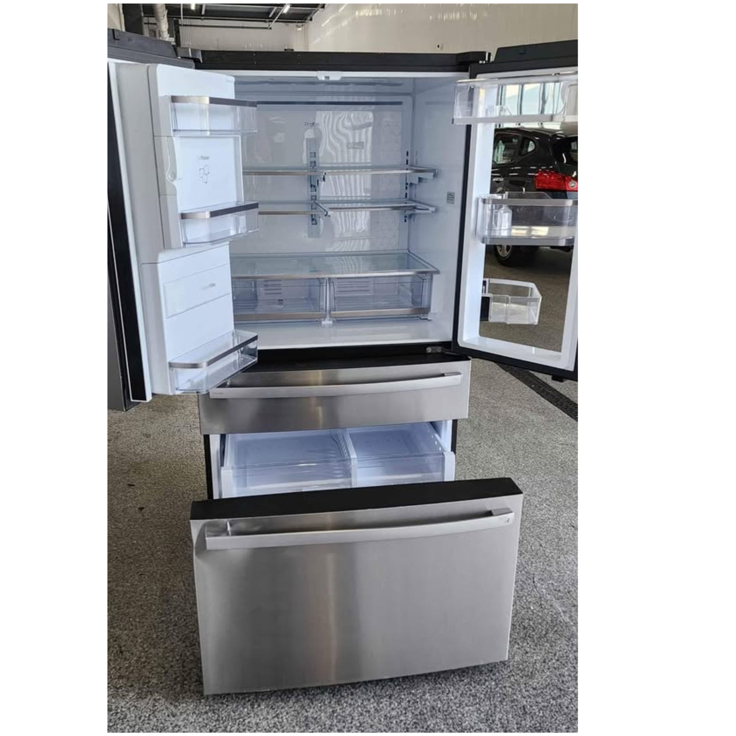 A stainless steel French door refrigerator with both doors open, showing the interior shelves and drawers, photographed in a showroom or warehouse setting.