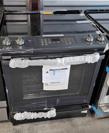 A black gas range with a glass cooktop and oven door, still wrapped in protective plastic and a paper label hanging on the door, photographed in a showroom.