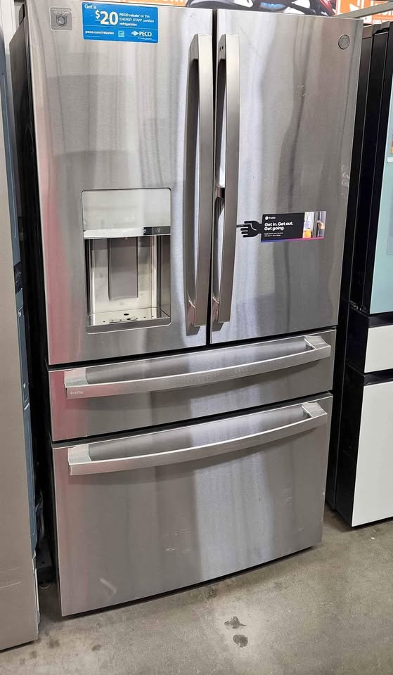 A stainless steel GE French door refrigerator with a bottom freezer drawer and a water and ice dispenser on the left door, photographed in a store setting.