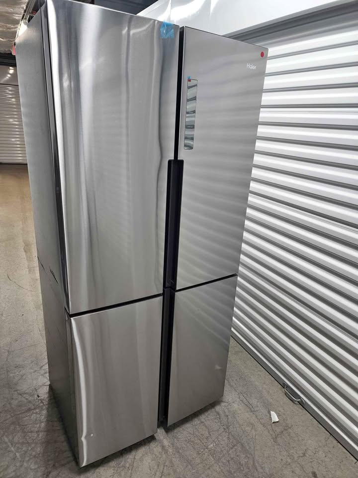 A stainless steel four-door refrigerator with a sleek brushed metal finish, standing in an industrial warehouse setting with a corrugated metal door and concrete floor.