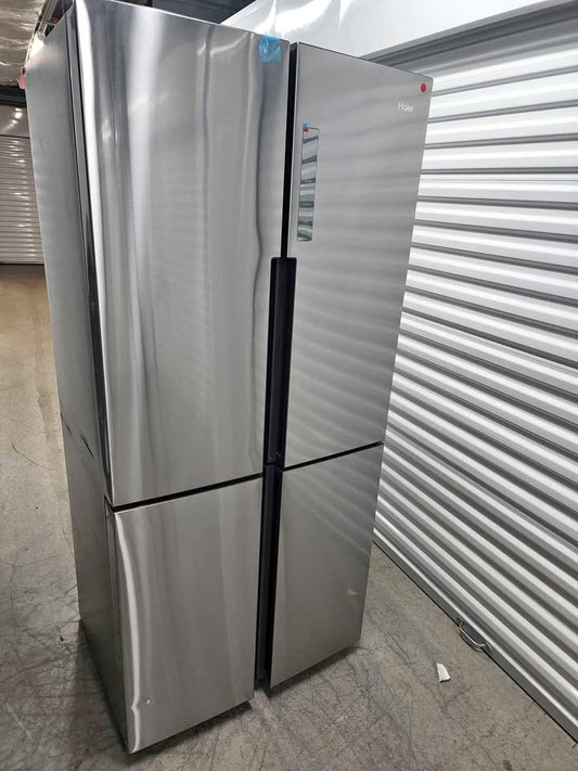 A stainless steel four-door refrigerator with a sleek brushed metal finish, standing in an industrial warehouse setting with a corrugated metal door and concrete floor.
