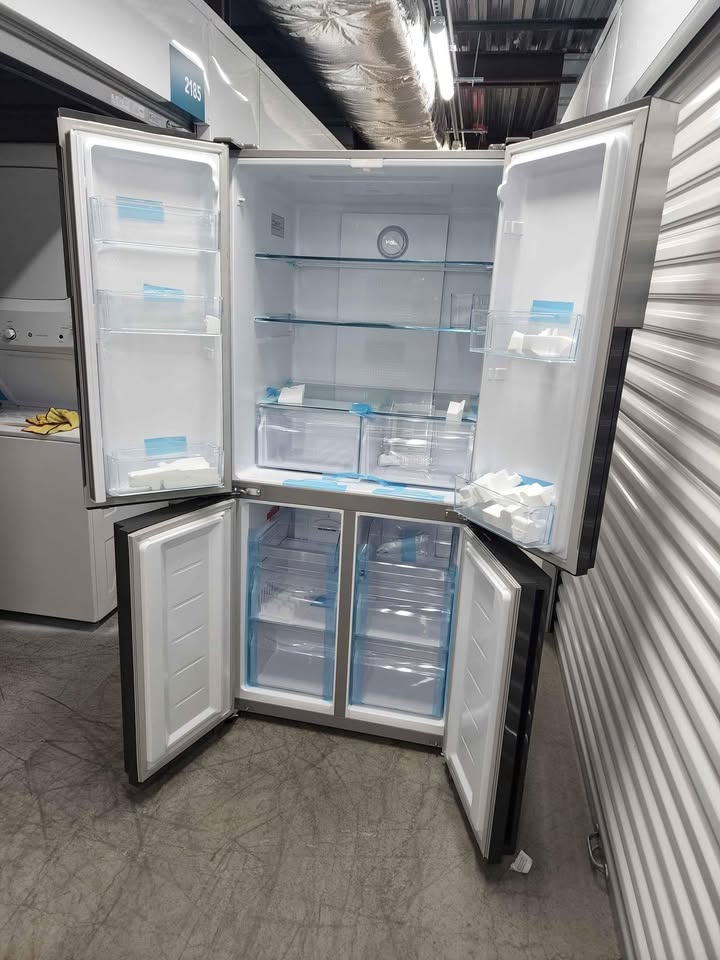 A stainless steel four-door refrigerator with all four doors open, showing the empty interior shelves, door bins, and lower transparent storage drawers in a warehouse or showroom setting. condition new