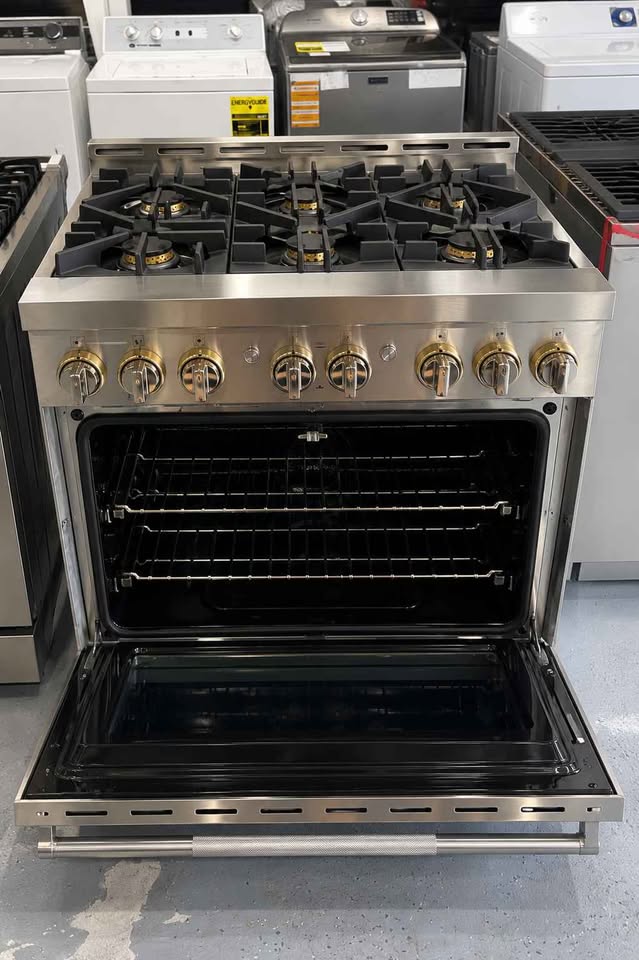 A stainless steel gas range with an open oven door, showing black interior racks and a black glass door, photographed in a showroom with other appliances in the background.