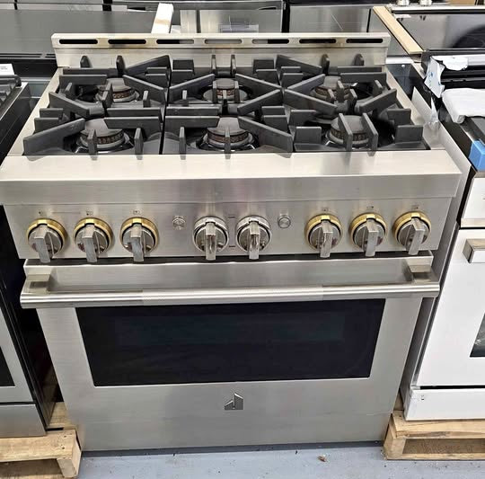 A stainless steel freestanding gas range with six front control knobs and a black oven door, photographed in a showroom or warehouse setting with other appliances visible around it.