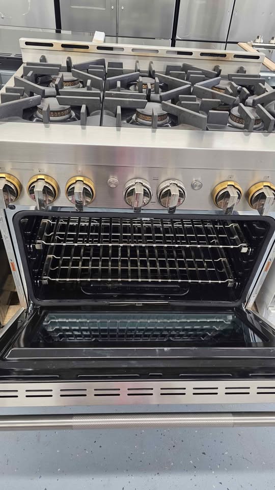 A stainless steel gas range with an open oven door showing interior racks, set against a kitchen or showroom background.