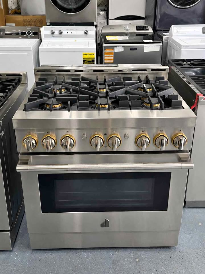 A stainless steel  gas range stove with six gold-toned control knobs and a black oven window, photographed in a showroom with other appliances in the background.