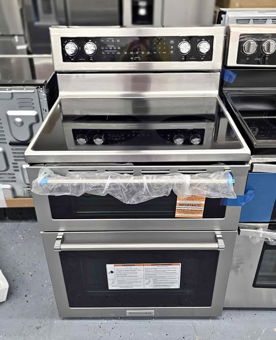 A stainless steel freestanding range with a black glass cooktop, five burners, and an underoven drawer, photographed in a showroom with other appliances in the background.