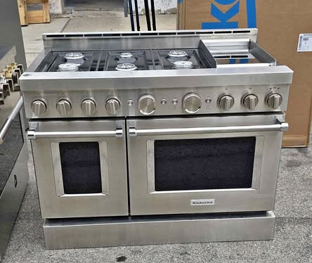 A stainless steel KitchenAid 48" Commercial-Style Gas Range with Griddle, freestanding gas range with multiple front knobs and two oven doors, photographed in a warehouse or showroom setting with a large cardboard box behind it.