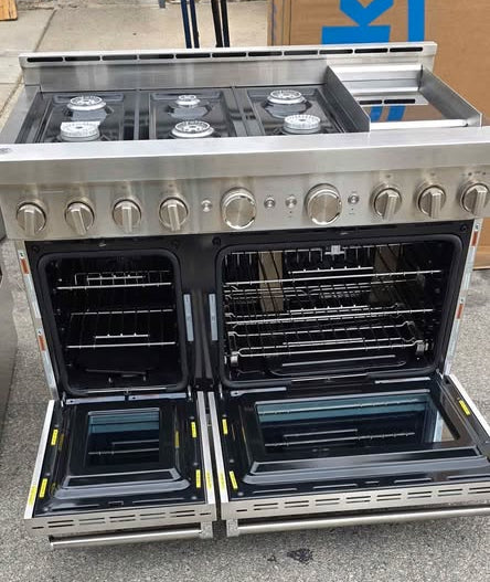 A stainless steel gas range with two open ovens and a built-in cooktop, photographed outdoors on a concrete surface with store boxes in the background.