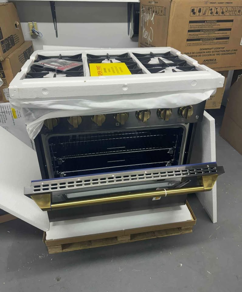 A freestanding gas range stove with an open oven door, wrapped in protective plastic on top, photographed in a warehouse or storage area with cardboard boxes around it.