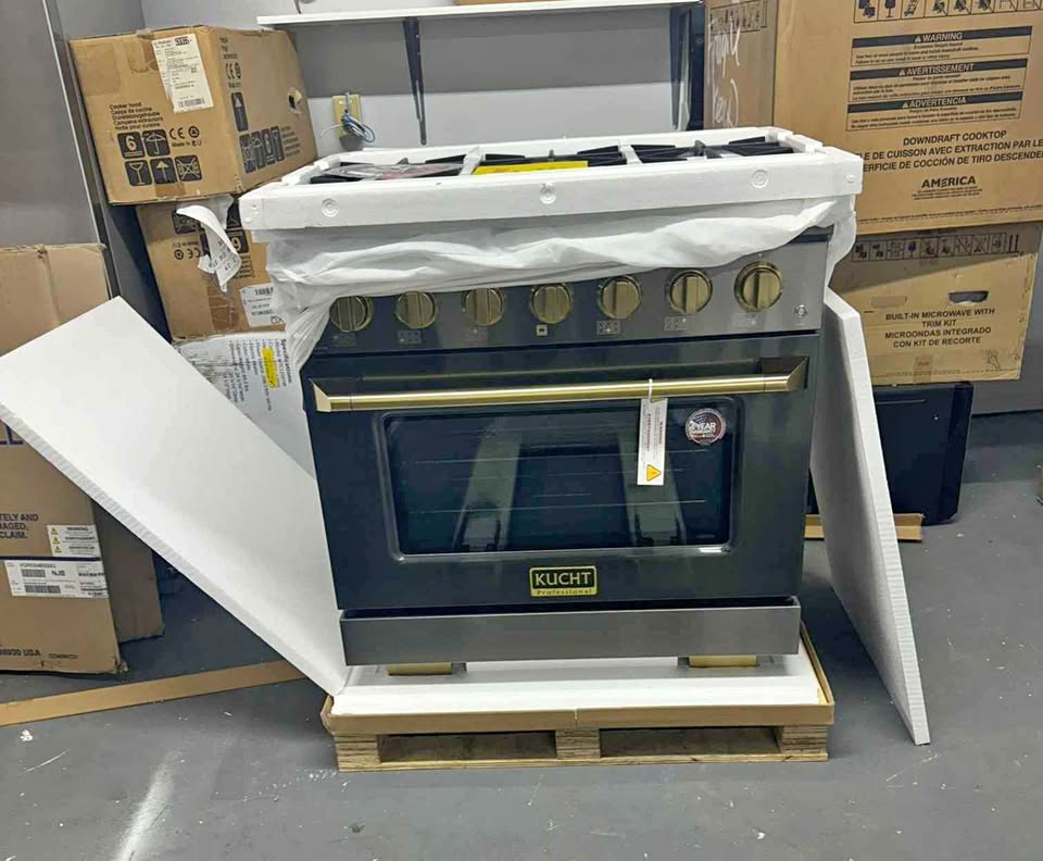 A Kuhnitz freestanding gas range oven with a stainless steel front and gold-tone knobs, sitting on a wooden pallet with protective white foam wrapping still attached. Cardboard boxes are stacked in the background on a warehouse floor.