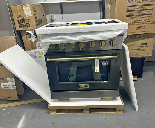 A Kuhnitz freestanding gas range oven with a stainless steel front and gold-tone knobs, sitting on a wooden pallet with protective white foam wrapping still attached. Cardboard boxes are stacked in the background on a warehouse floor.