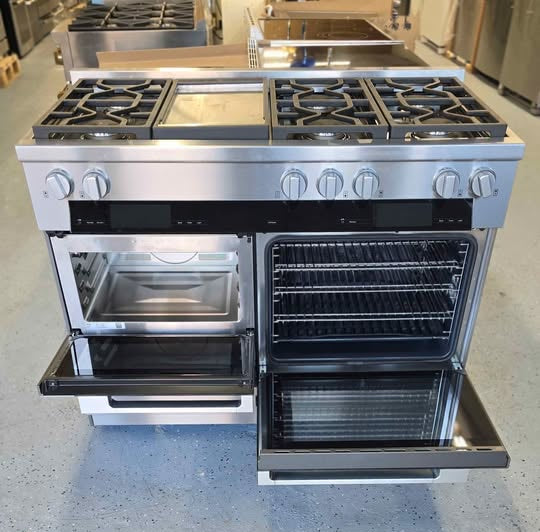 A stainless steel freestanding gas range with two open ovens, photographed in a showroom or warehouse setting on a speckled gray floor.