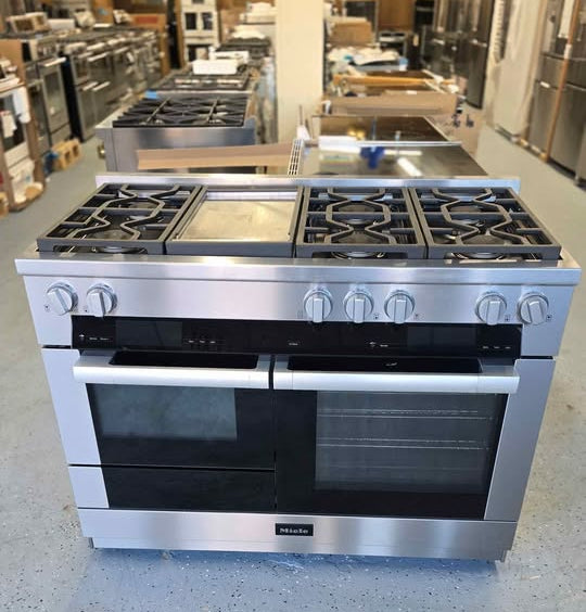 A stainless steel freestanding gas range with black glass oven doors and control knobs, photographed in a showroom or warehouse setting with other appliances in the background.