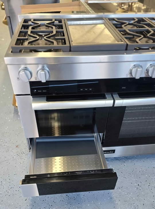 A stainless steel gas range with a black glass oven door and an open lower drawer, photographed in a showroom or warehouse setting on a speckled gray floor.