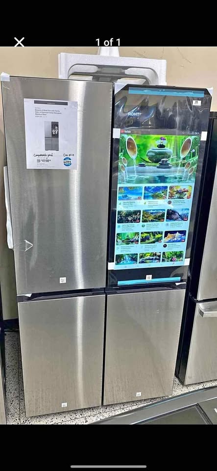 A stainless steel refrigerator with a large vertical touchscreen display showing colorful aquarium-style images, photographed in a showroom. A product info sheet is attached to the upper left door, and other appliances are partially visible around it.