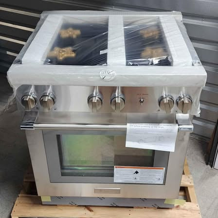 A stainless steel commercial range hood or ventilation unit with multiple control knobs, wrapped in protective plastic, sitting on a wooden pallet against a corrugated metal wall.