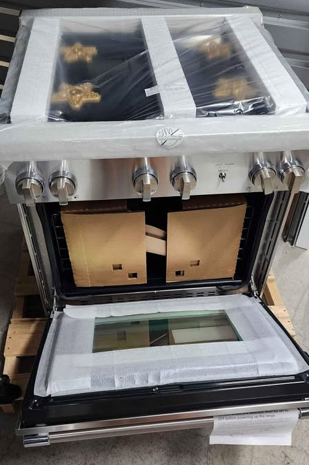 A stainless steel gas range with an open oven door, wrapped in protective plastic on the top surface and drawers, photographed on a wooden pallet in a warehouse setting.