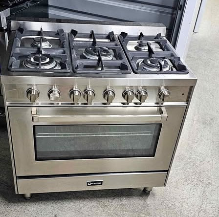 A stainless steel freestanding gas range, Verona 36" Freestanding Pro-Style Gas Range with five front burners, a large oven window, and multiple control knobs, photographed indoors on a concrete floor in front of a gray slatted wall.