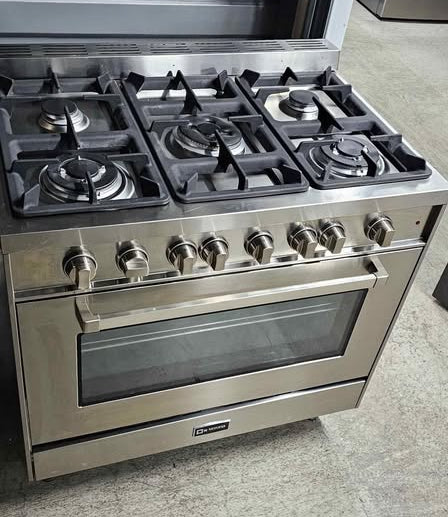 A stainless steel freestanding gas range with five burners, a large oven window, and multiple control knobs, photographed in what appears to be a showroom or warehouse setting.