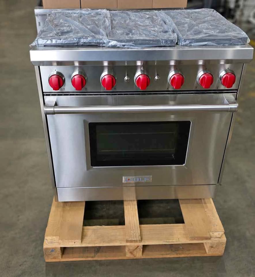 A stainless steel freestanding gas range with six red control knobs and a black oven window, sitting on a wooden pallet in a warehouse setting.