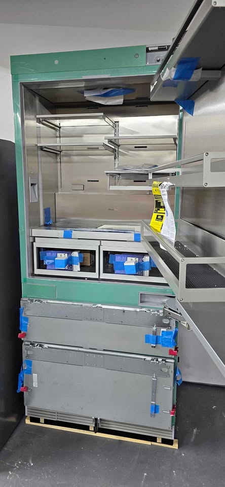 A large industrial stainless steel storage cabinet or rack system with a green outer frame, open upper door, shelving inside, and two stacked gray metal modules on a wooden base. Blue tape and a yellow tag are visible, and the unit is photographed in a warehouse-like setting.